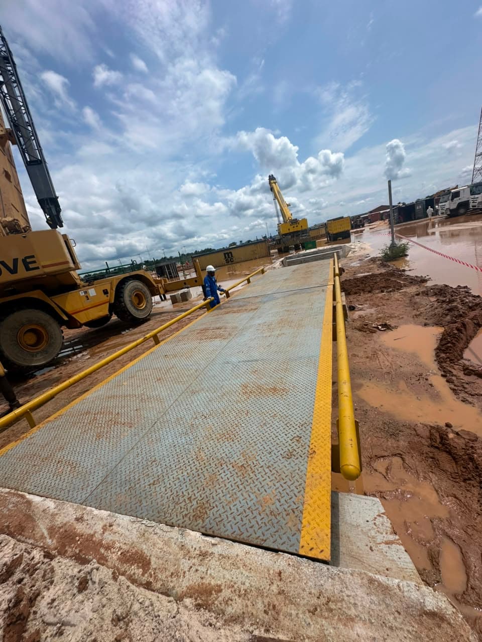 Newly installed steel weighbridge deck with yellow guardrails at a container yard, cranes in the background